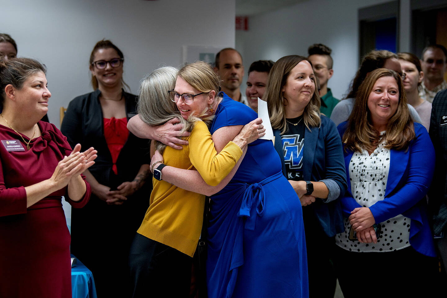 Two individuals embrace warmly in the center of a room where others watch and clap, suggesting a celebratory or emotional moment.